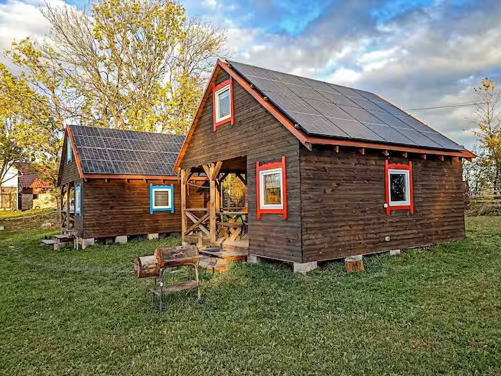 Two rustic wooden cabins are situated on a grassy area, featuring colorful window frames and solar panels on the roofs. A welcoming porch with seating is visible in front of each cabin, surrounded by trees and an open sky.
