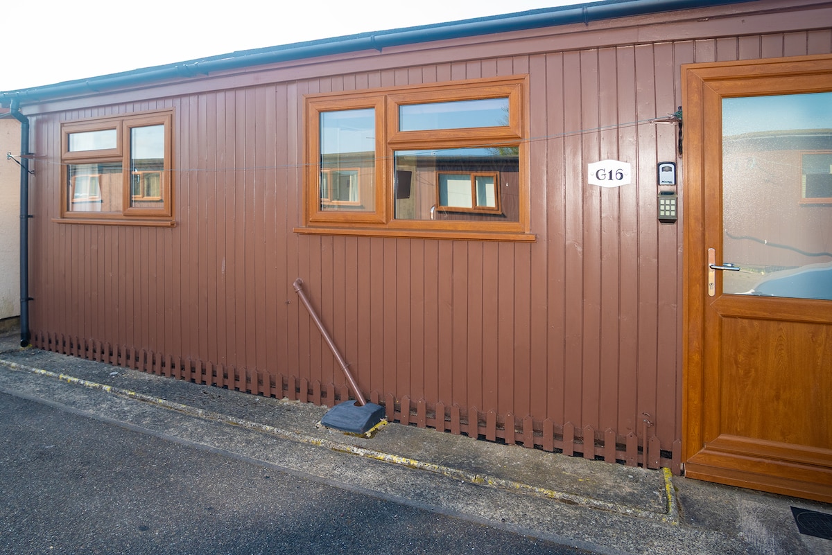 A single-story building is shown, constructed with a brown panel exterior. The front entrance features a wooden door with a metal number plate. Two large windows provide natural light, framed in matching wood, while a broom leans against the side.