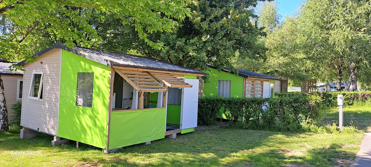 Two Tithomes are visible, featuring modern designs with vibrant green accents. Each unit is elevated slightly above the ground on sturdy supports, and sheltered porches are highlighted. The grassy area is adorned with trees, providing a natural environment for relaxation.