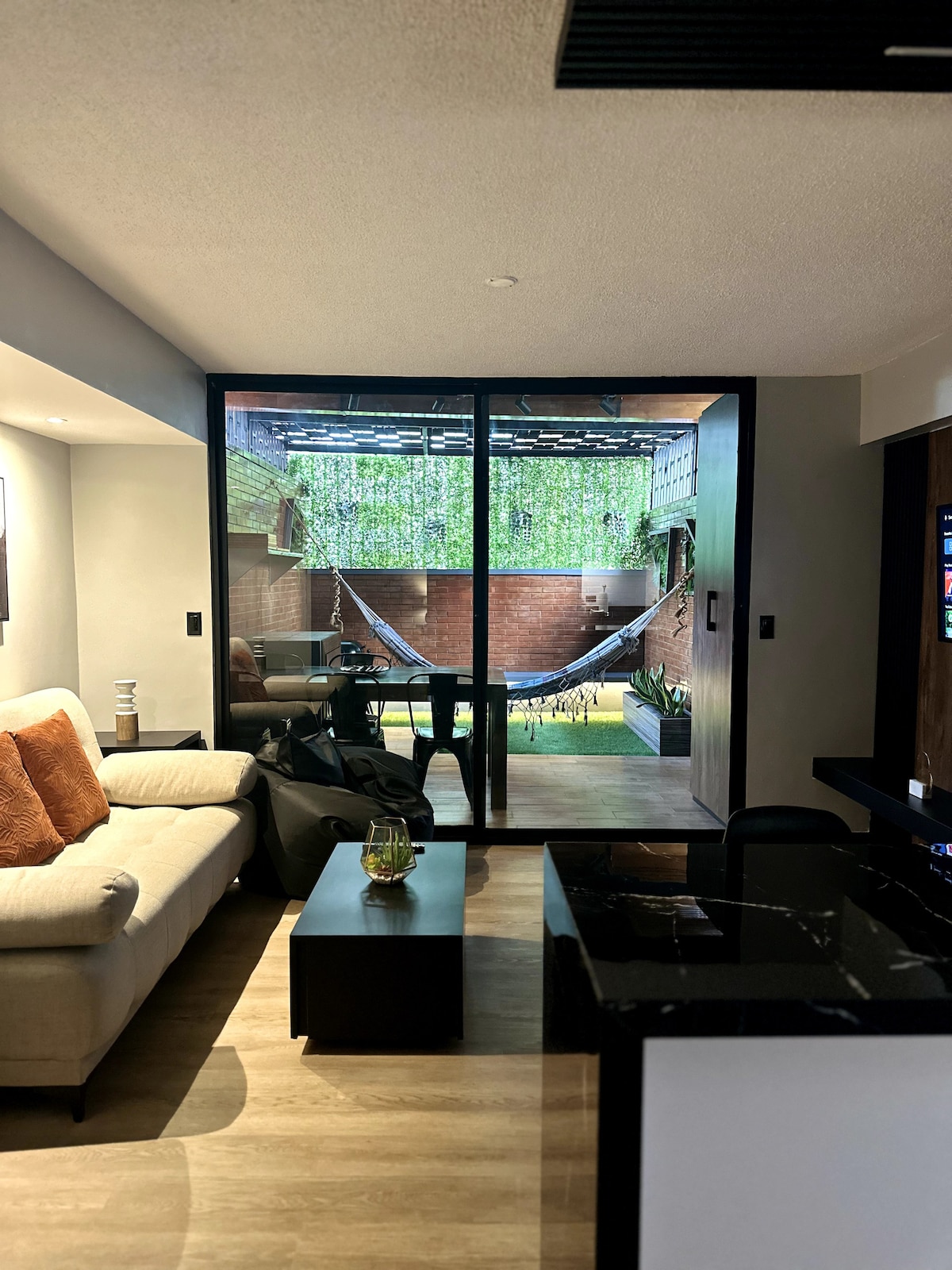 A living area is shown with a comfortable sofa, accent pillows, and a dark coffee table. Large glass doors open to a private terrace featuring a hammock and green plants, creating a seamless connection between the indoor and outdoor spaces.