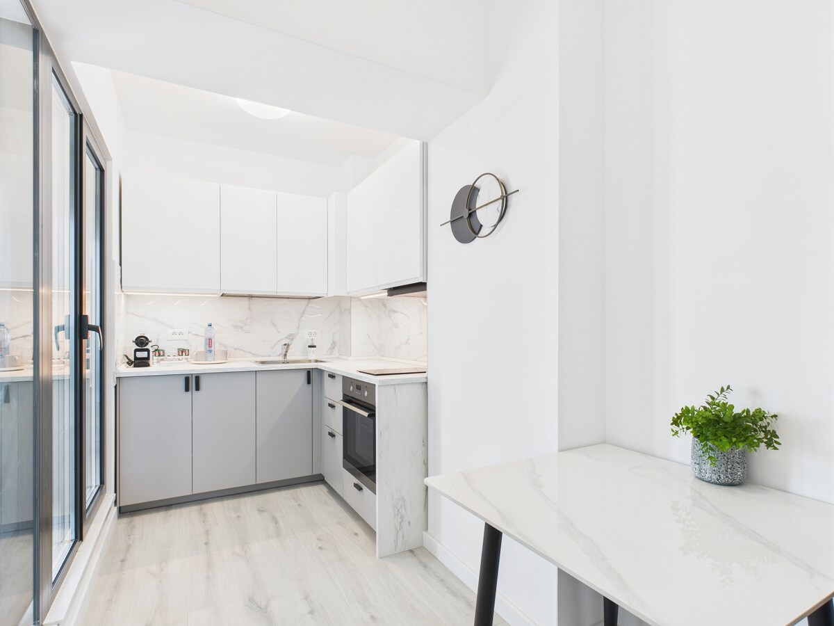 A modern kitchen is showcased with sleek white cabinets and gray accents. Stainless steel appliances, including an oven and stove, are positioned along the wall. Natural light floods the space through large glass doors, enhancing the bright and airy atmosphere.