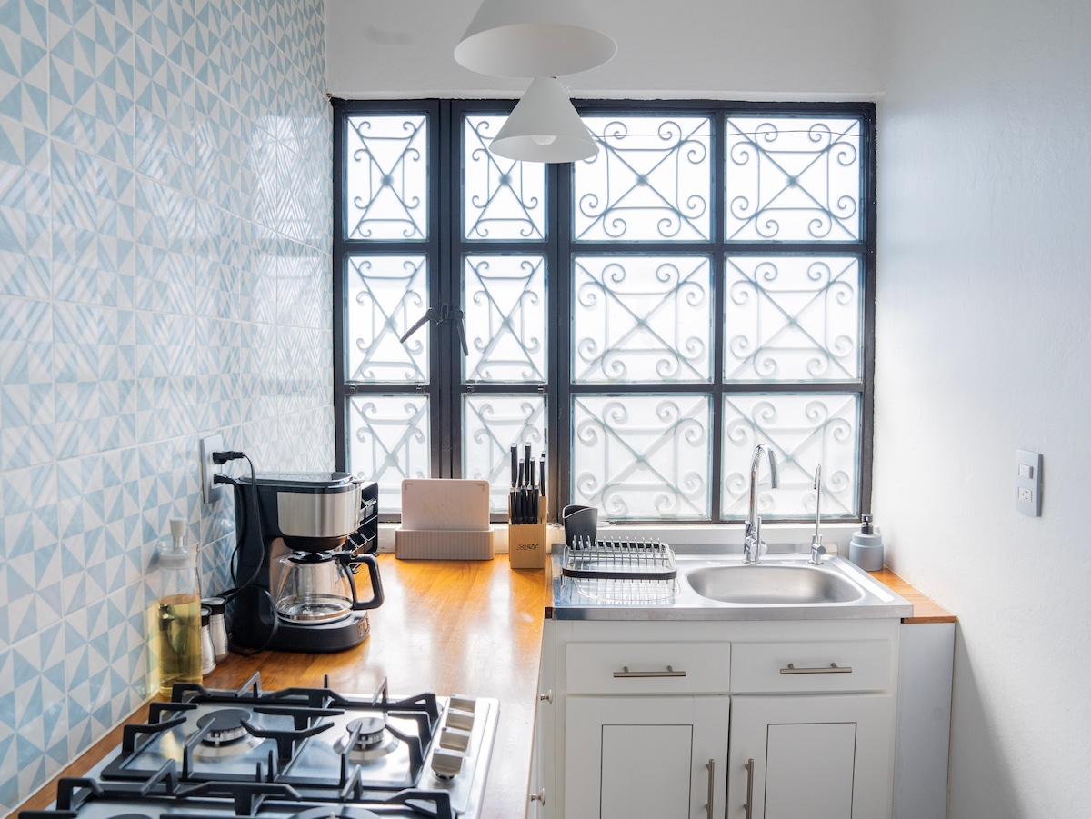 A modern kitchen showcases a sleek countertop and a gas stove. Natural light fills the space through large, wrought-iron framed windows. Kitchen essentials, including a coffee maker and dish rack, are organized neatly. The tiled backsplash features a geometric pattern that adds a stylish touch.
