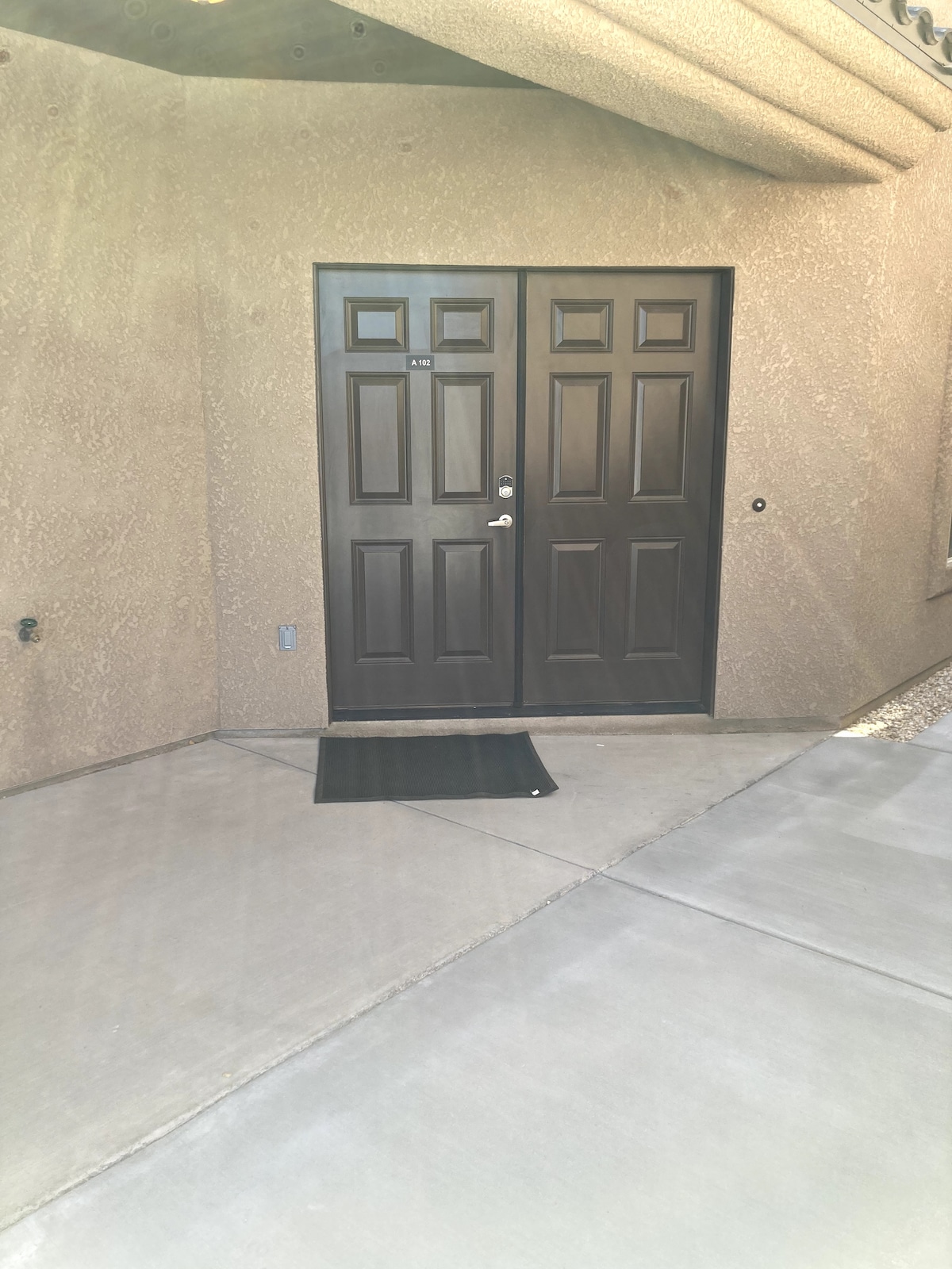 The entrance features a pair of dark double doors positioned against a textured beige wall. A small black mat lies on the concrete pathway leading to the doors, offering a welcoming touch. Natural light casts soft shadows, enhancing the simplicity of the entryway.