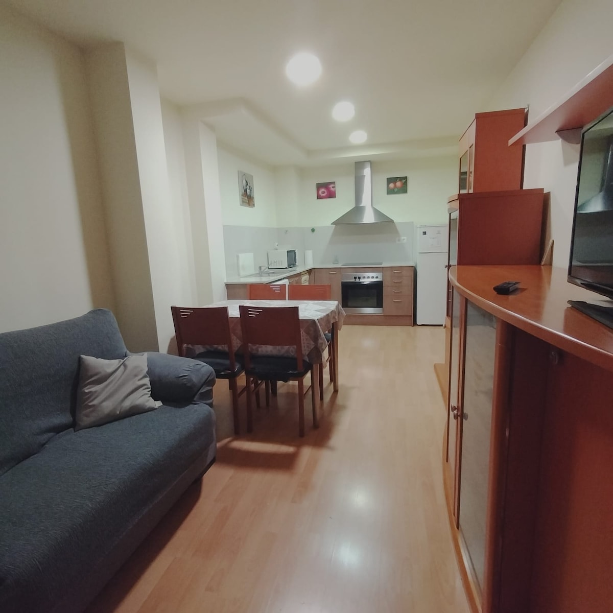 An open-plan kitchen and dining area is shown, featuring a modern stovetop and hood. A dining table with four chairs is positioned next to a comfortable sofa. Natural light fills the space, highlighting the wooden cabinets and light-colored flooring.
