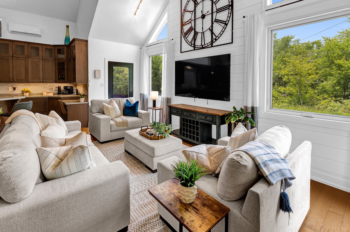 A bright and airy living area features two comfortable sofas arranged around a wooden coffee table, with a large flat-screen TV mounted on the wall. Sunlight streams through multiple windows, illuminating the light-colored decor and accent plants, creating a relaxed space.