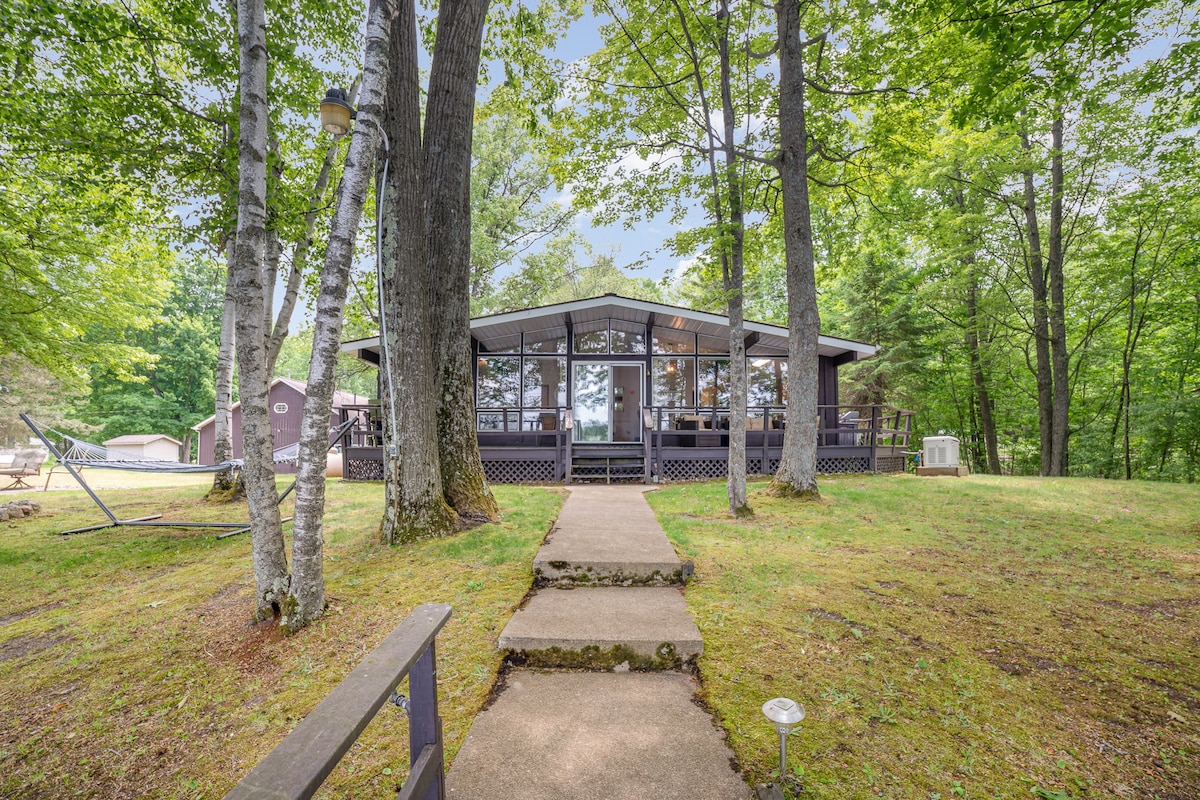 A serene lodge is situated among lush greenery, featuring large glass windows that offer views of the surrounding nature. A stone pathway leads to the entrance, bordered by tall trees. A spacious wrap-around deck extends from the house, providing outdoor living space.