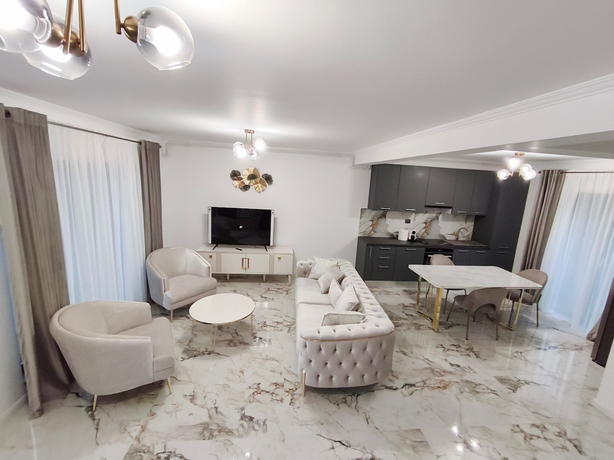 A spacious living area showcases elegant furniture, including plush armchairs and a tufted sofa, arranged around a round coffee table. A large dining table sits nearby. Natural light floods in through sheer curtains, while stylish light fixtures add a modern touch to the design.