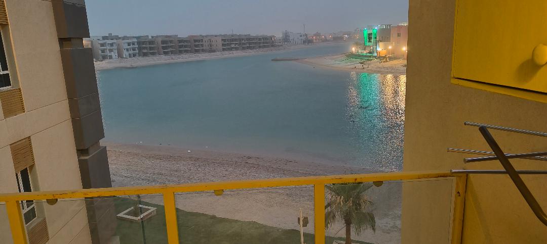 A balcony view showcases a calm waterway bordered by sandy shores, with distant buildings lining the bank. Subtle reflections of lights can be seen dancing on the water's surface, while palm trees are partially visible in the foreground, adding a touch of greenery to the scene.