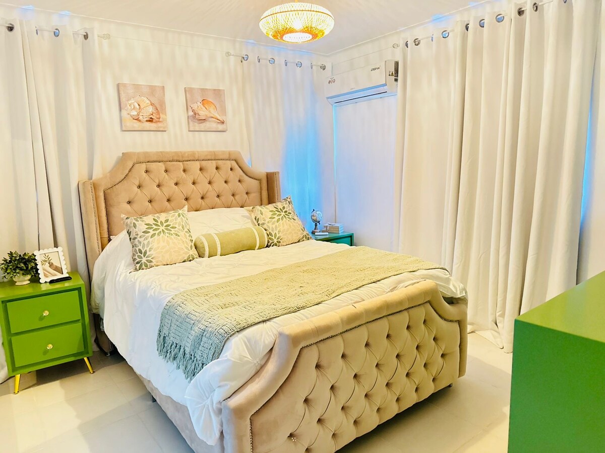 A well-appointed bedroom features a plush, tufted bed adorned with decorative pillows. Two bedside tables provide additional storage, and a soft green blanket accents the bed. Light curtains diffuse natural light, while a stylish ceiling fixture adds a modern touch.