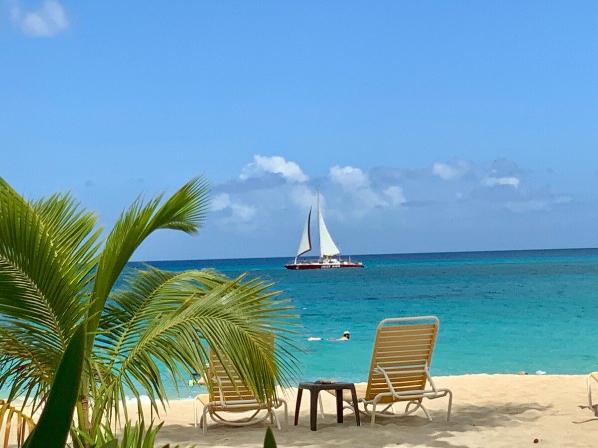 A serene beach scene features two lounge chairs positioned on soft sand, overlooking turquoise waters. A sailboat with white sails glides in the distance under a clear blue sky, while fluffy white clouds add a tranquil touch to the horizon.