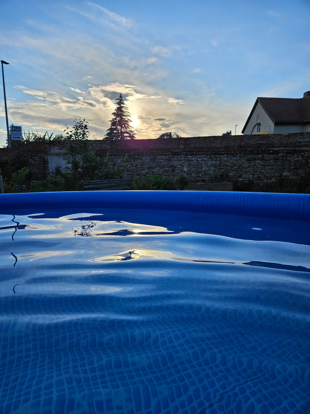 The image features a pool with calm, rippling water reflecting the sky. A sunset casts warm light over the scene, and a hint of greenery is visible around the pool's edge, creating a serene outdoor environment.
