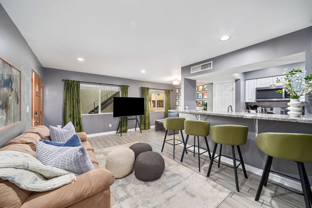 A spacious living area is highlighted by a comfortable sofa, adorned with decorative pillows. Several bar stools are arranged around a kitchen island under soft lighting. Large windows bring in natural light, creating an airy feel complemented by green curtains.