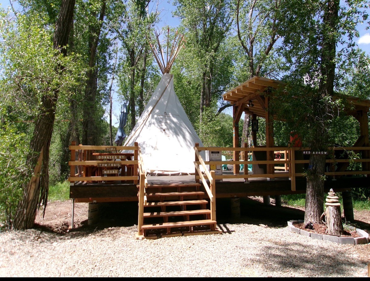 A spacious riverside tipi is set on a wooden deck surrounded by trees. A staircase leads up to the entrance, which is framed by a wooden railing. Natural light filters through the trees, creating a serene atmosphere in the outdoor setting.
