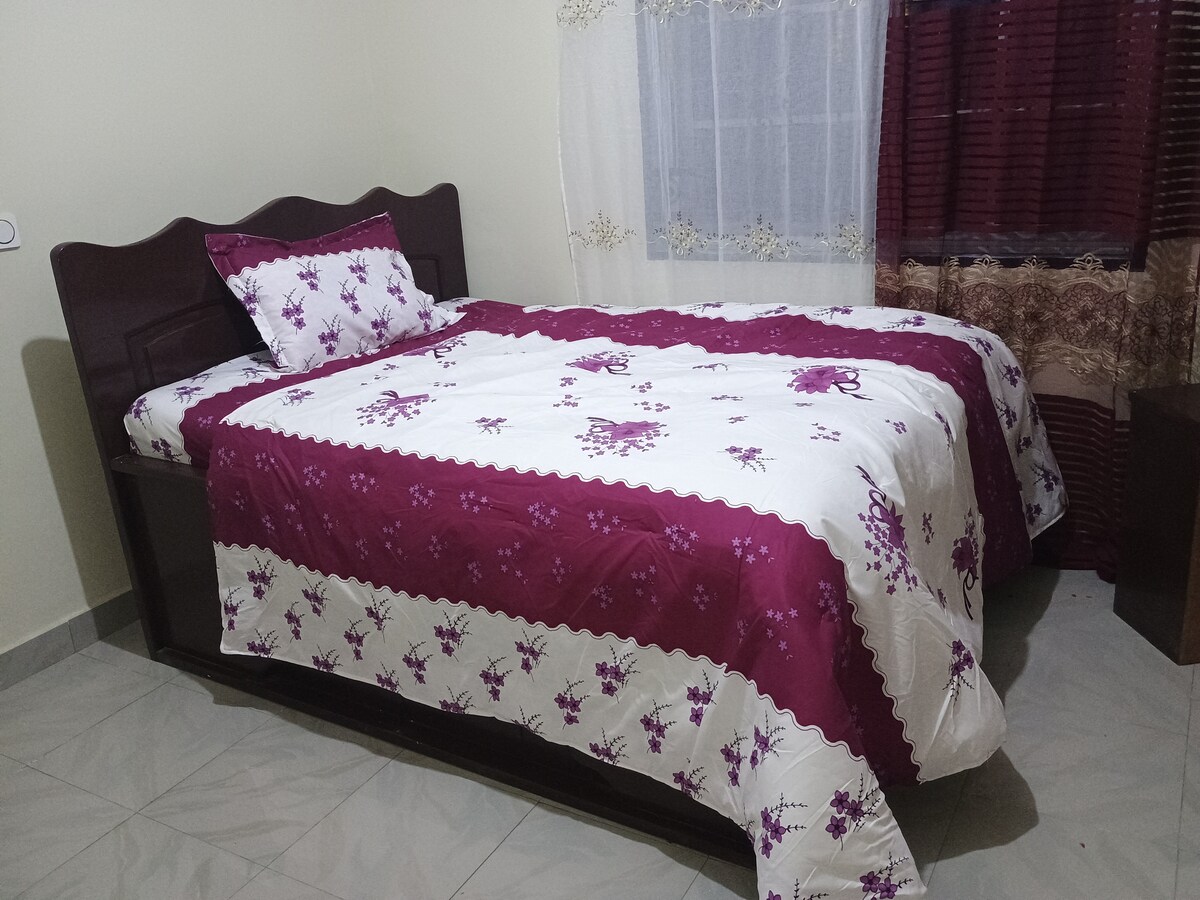 A well-made bed is presented with floral-patterned bedding in shades of purple and white. A decorative pillow rests at the head of the bed. Natural light filters through a nearby window, which is adorned with sheer and patterned curtains.