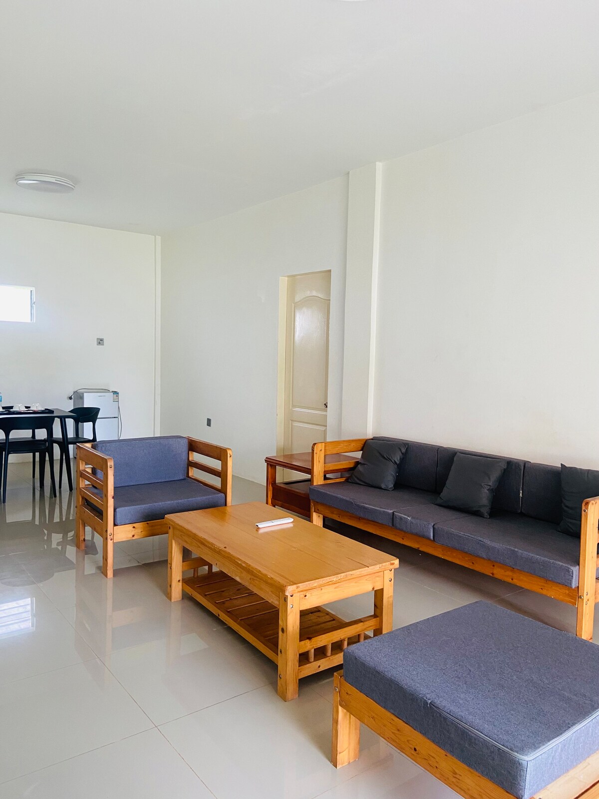 A living area is visible, showcasing a combination of wooden seating with cushioned gray upholstery. A coffee table sits centrally, complementing the arrangement. Light walls and reflective flooring create an open and airy ambiance, while a small dining area can be seen in the background.