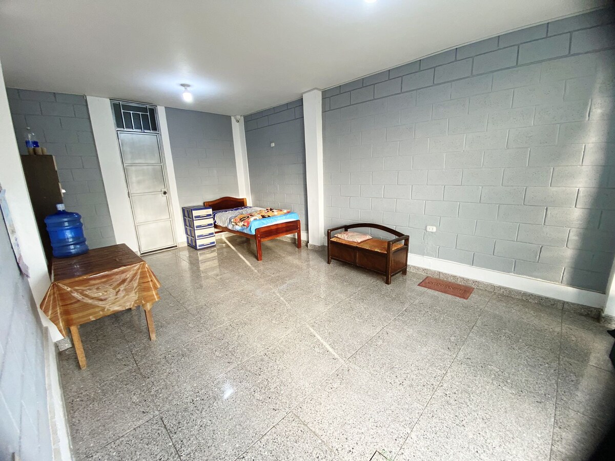 A spacious room is shown with smooth tiled flooring and gray walls. Two beds, one with a colorful blanket, are positioned in the space alongside a wooden table and a seating area. A water dispenser is visible near the entrance.