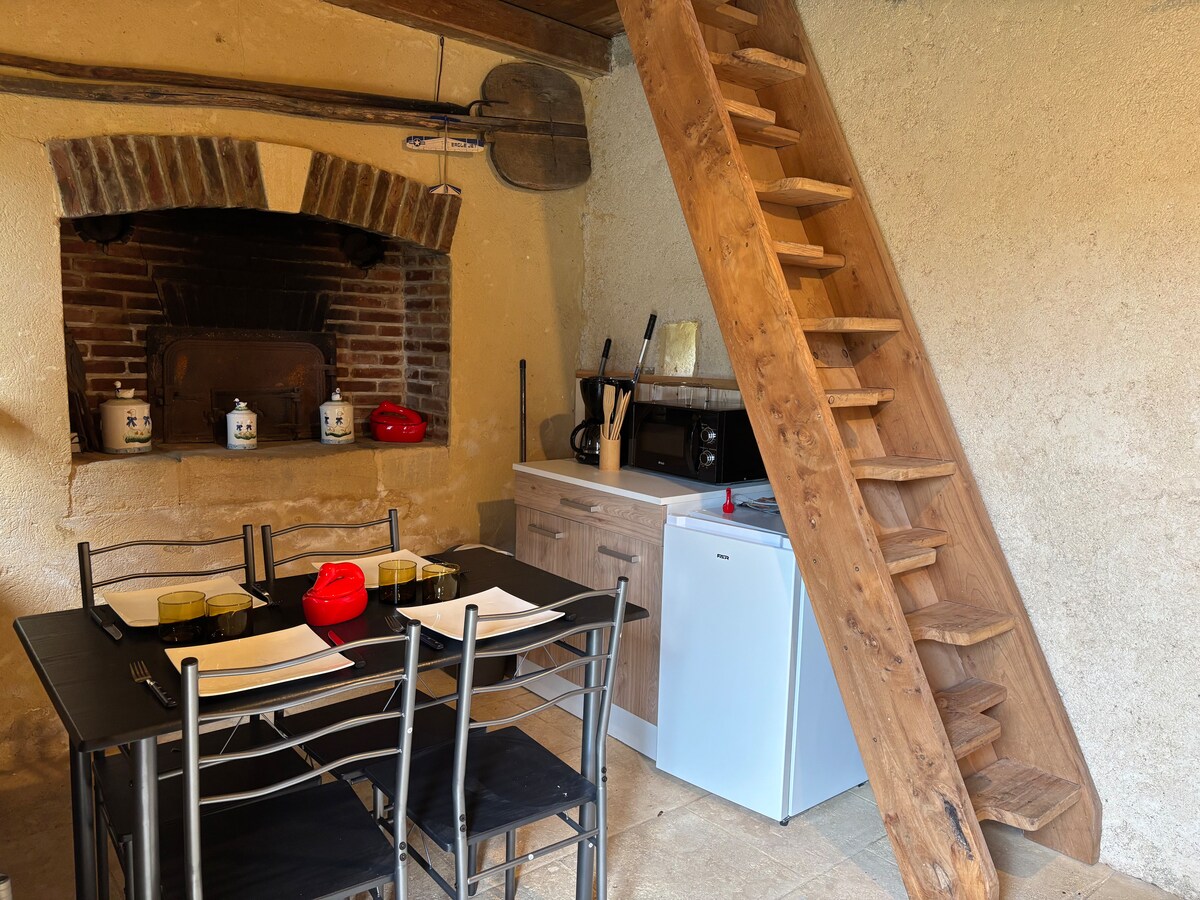An open kitchenette area is highlighted by exposed wooden beams and a decorative stone fireplace. A dining table set for four is visible, accompanied by black chairs. A compact refrigerator and a kitchenette are situated along the wall, with stairs leading to the upper level.