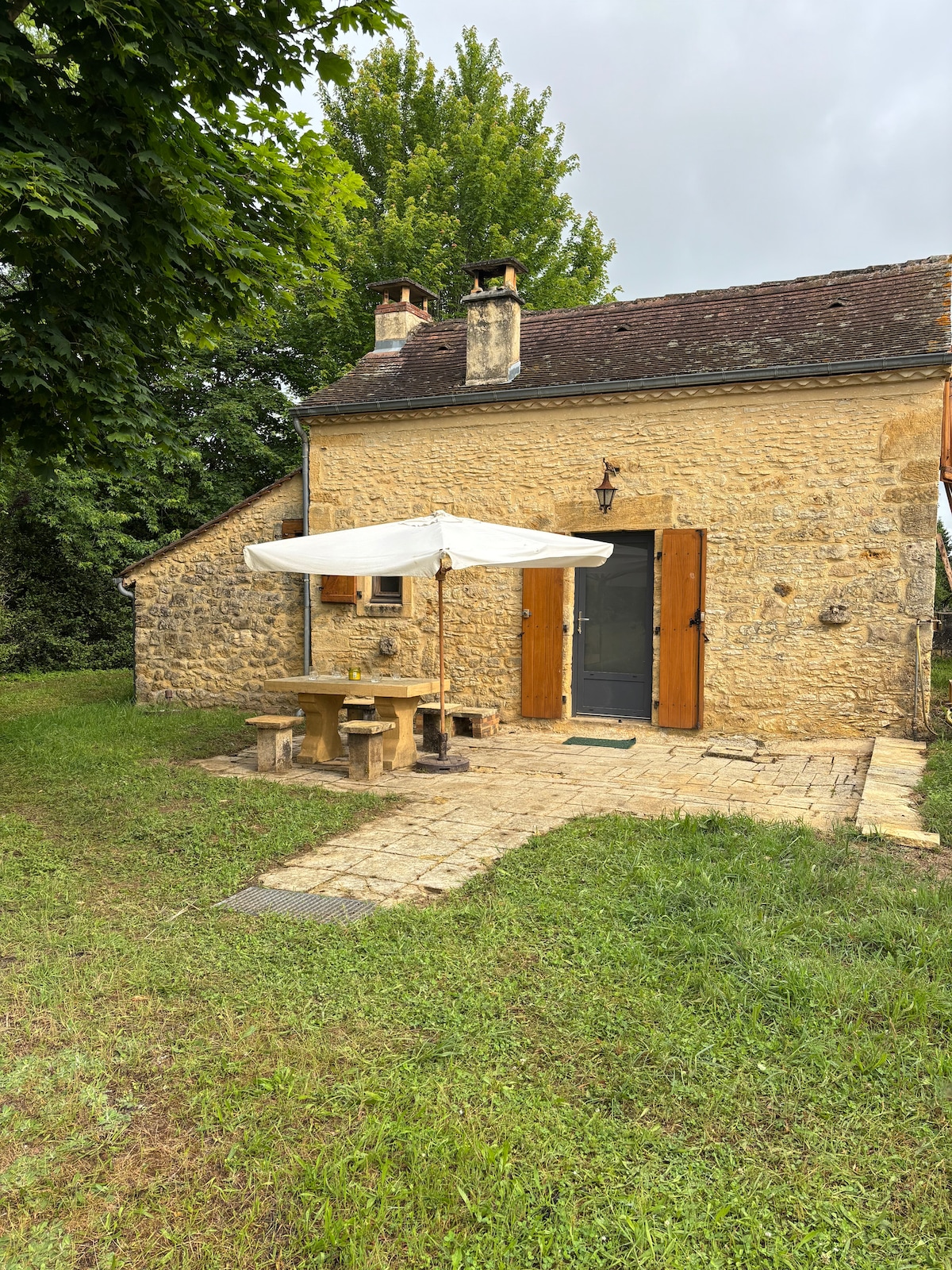 A rustic stone cottage is showcased, featuring a light-colored facade with timber accents. A wooden table is placed under a white umbrella, surrounded by a grassy area. The entrance is framed by two doors, inviting guests into the cozy interior.