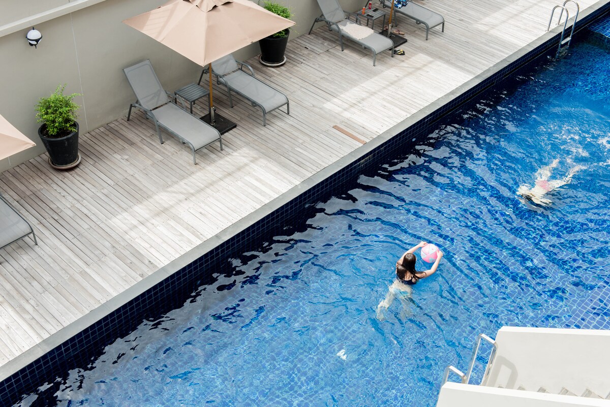A serene outdoor swimming pool is surrounded by wooden decking and complemented by lounge chairs and shaded umbrellas. Two individuals can be seen enjoying the water, with the clear blue surface reflecting sunlight, creating a relaxing environment for guests.