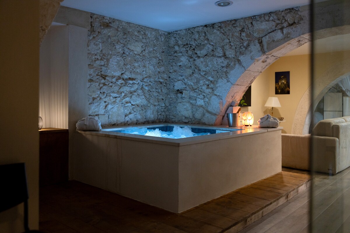 A private hydromassage pool is situated in a softly lit area, surrounded by textured stone walls. A cozy ambiance is created with gentle lighting and simple furnishings, while two neatly folded towels are placed at the pool's edge, inviting relaxation.