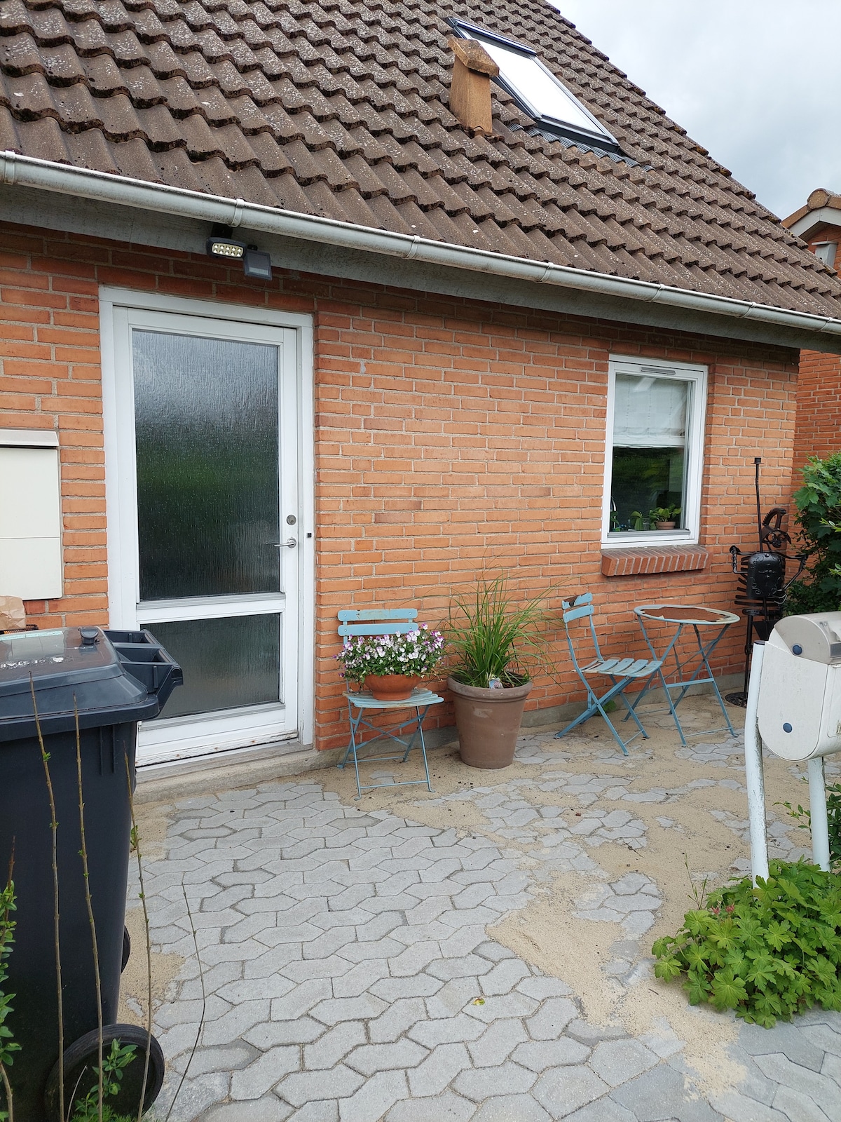 The exterior of a brick house displays a welcoming front entrance with a glass door. Two light blue chairs surround a small table, accompanied by potted plants. A trash bin is visible, along with a mailbox positioned nearby, adding to the home's practical features.