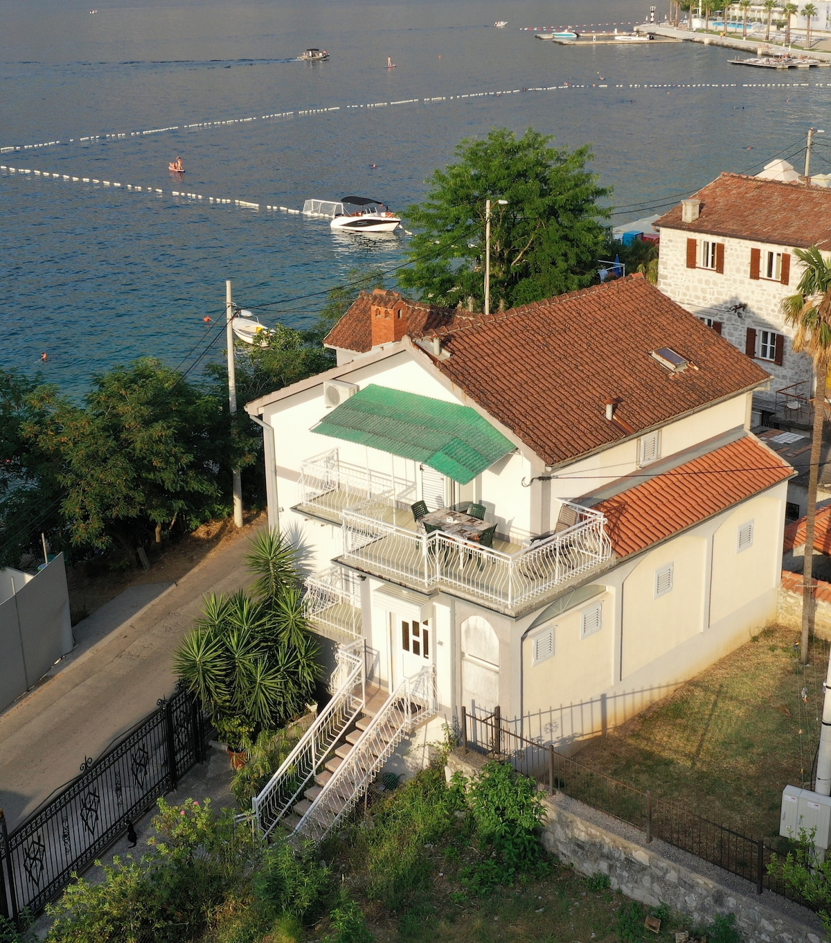 A two-story house is positioned just meters from the water, featuring a green awning over a balcony. The exterior exhibits a combination of stone and stucco, surrounded by trees and a driveway. Boats can be seen in the water nearby, enhancing the coastal setting.