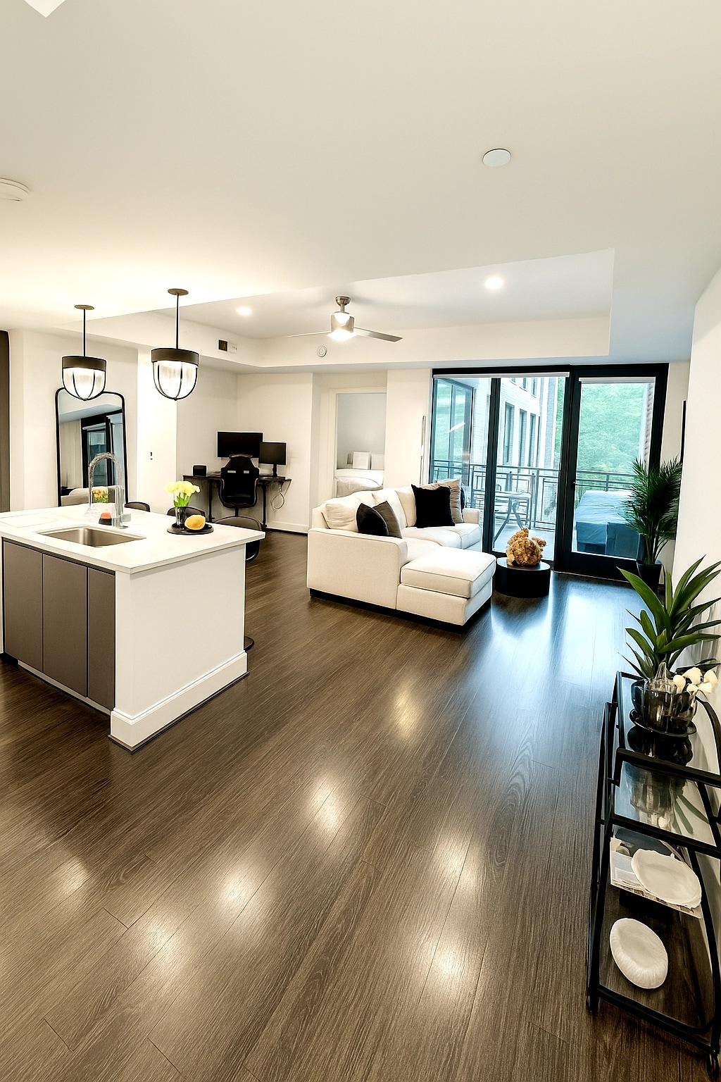 A modern living area is displayed, featuring a light-colored sectional sofa with dark accent pillows. A sleek kitchen island with bar seating is visible, along with a workspace area. Large glass doors provide access to an outdoor balcony, allowing natural light to fill the space.