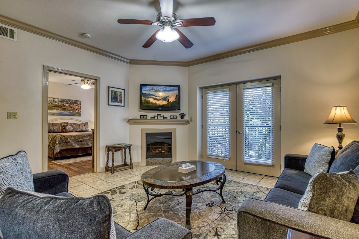 A comfortable living area features a dark gray couch and matching armchairs arranged around a circular table. A fireplace sits beneath a mounted television. Two sets of glass doors open to a serene outdoor space, while soft lighting from a nearby lamp enhances the environment.