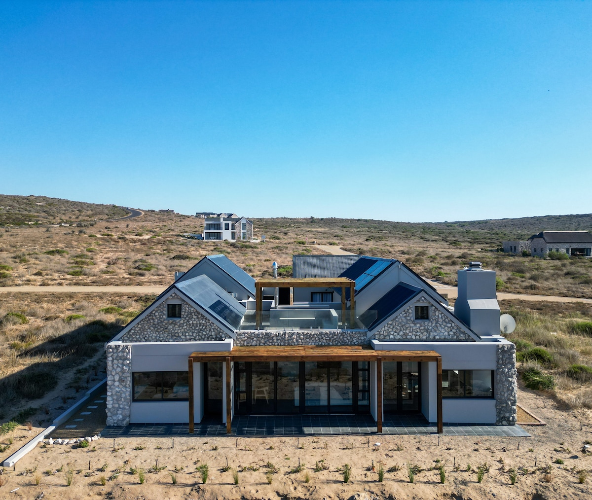 A spacious coastal retreat is showcased from an aerial perspective, revealing a contemporary design with a combination of stone and sleek roofing materials. Surrounding vegetation is visible, highlighting the natural landscape. Large windows provide expansive views, inviting light into the interior spaces.