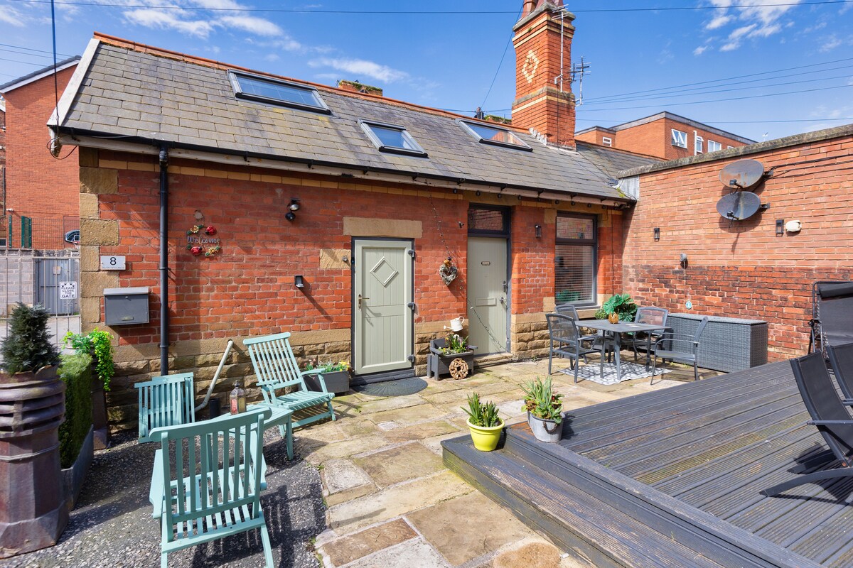 A shared secure courtyard is showcased with a combination of paved stones and wooden decking. Outdoor seating is provided with green chairs and a dining table. Planters with greenery add a natural touch to the space, complemented by the brick walls and historic clock tower in the background.