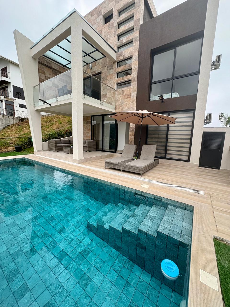 A modern home is depicted, featuring a private pool with clear water. Lounge chairs are positioned beside the pool under a large umbrella. The structure includes large glass windows and a spacious balcony, providing views of the surrounding area.
