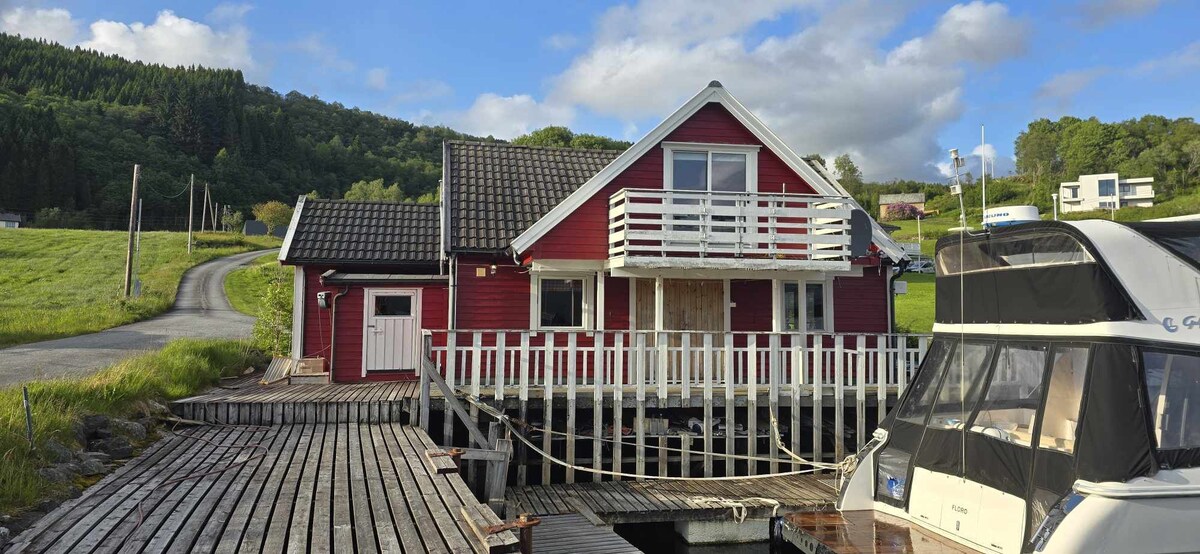 A red wooden house is located by the water, featuring a balcony and a covered entrance. A dock extends from the house into the water, accommodating a boat. Lush green hills rise in the background under a partly cloudy sky.