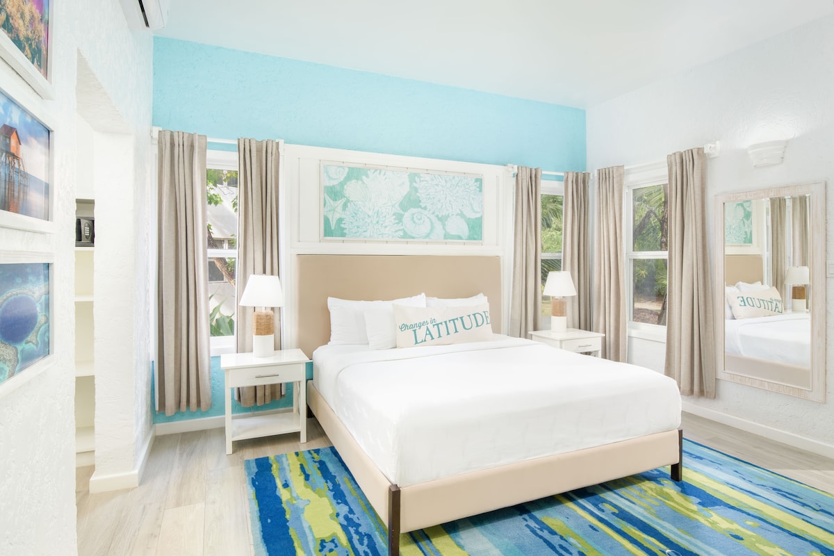 A bright and airy bedroom features a spacious bed with a light-colored headboard. Light blue walls and sheer curtains enhance the natural light. Decorative artwork and a large mirror add depth. A colorful area rug in shades of blue and green complements the space.