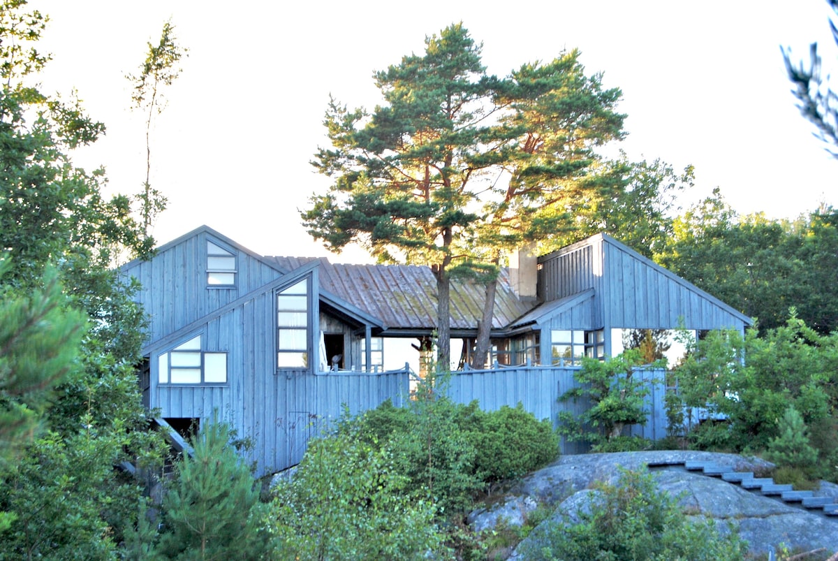 An architectural summer house is set amid a coastal forest, featuring a blend of wood and glass. The structure is elevated on rock, with a unique roofline and large windows that provide views of the surrounding nature.