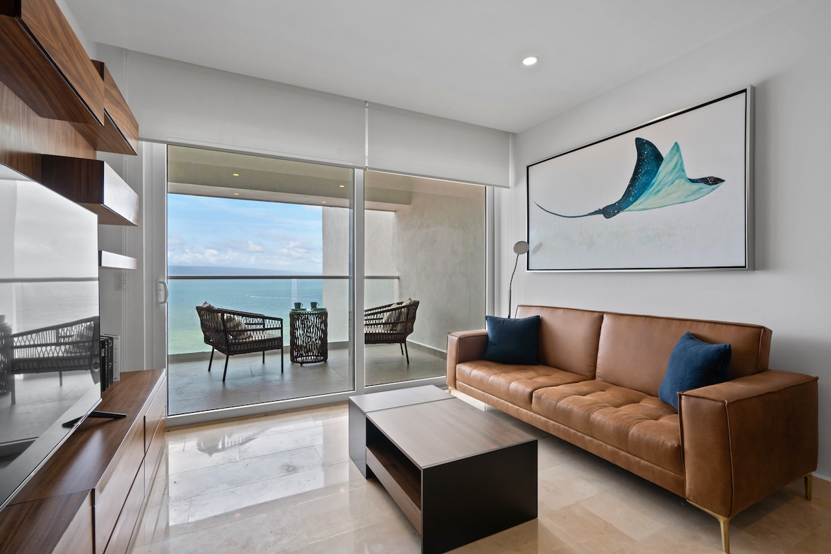 A modern living area features a brown leather sofa with blue accent pillows, complemented by a minimalist coffee table. Large glass doors open to a balcony, where two chairs are positioned to overlook the ocean. An abstract artwork depicting a manta ray adorns the wall.