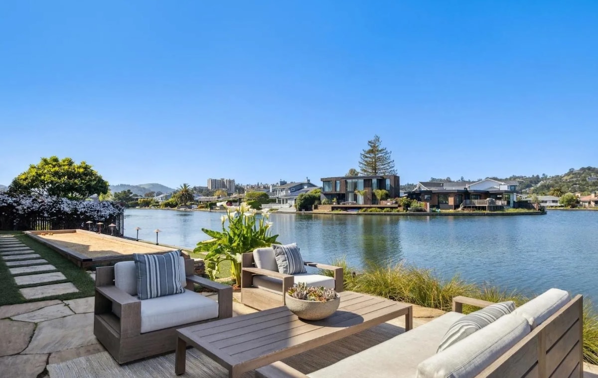 An outdoor patio features comfortable seating arrangements, overlooking a serene lagoon. Lush greenery and decorative plants punctuate the space, while a pathway leads toward the water. Scenic views of neighboring homes and the surrounding landscape are visible under a clear blue sky.