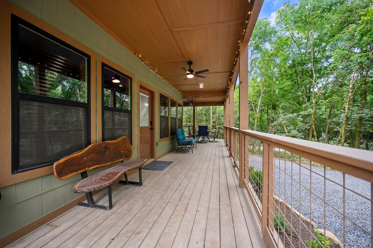 A spacious covered deck features wooden flooring and is framed by natural greenery. Comfortable seating options include a lounge chair and a rustic wooden bench. Soft outdoor lighting enhances the inviting atmosphere, while a fan is mounted on the ceiling for comfort.