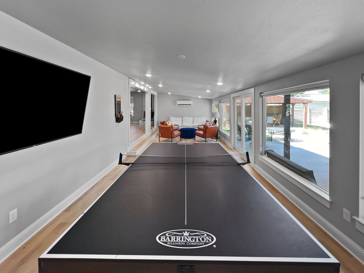 A game room is featured with a black ping pong table set in the foreground. Large windows allow natural light to fill the space, revealing a seating area with orange chairs and a white sofa beyond. A television is mounted on the wall.