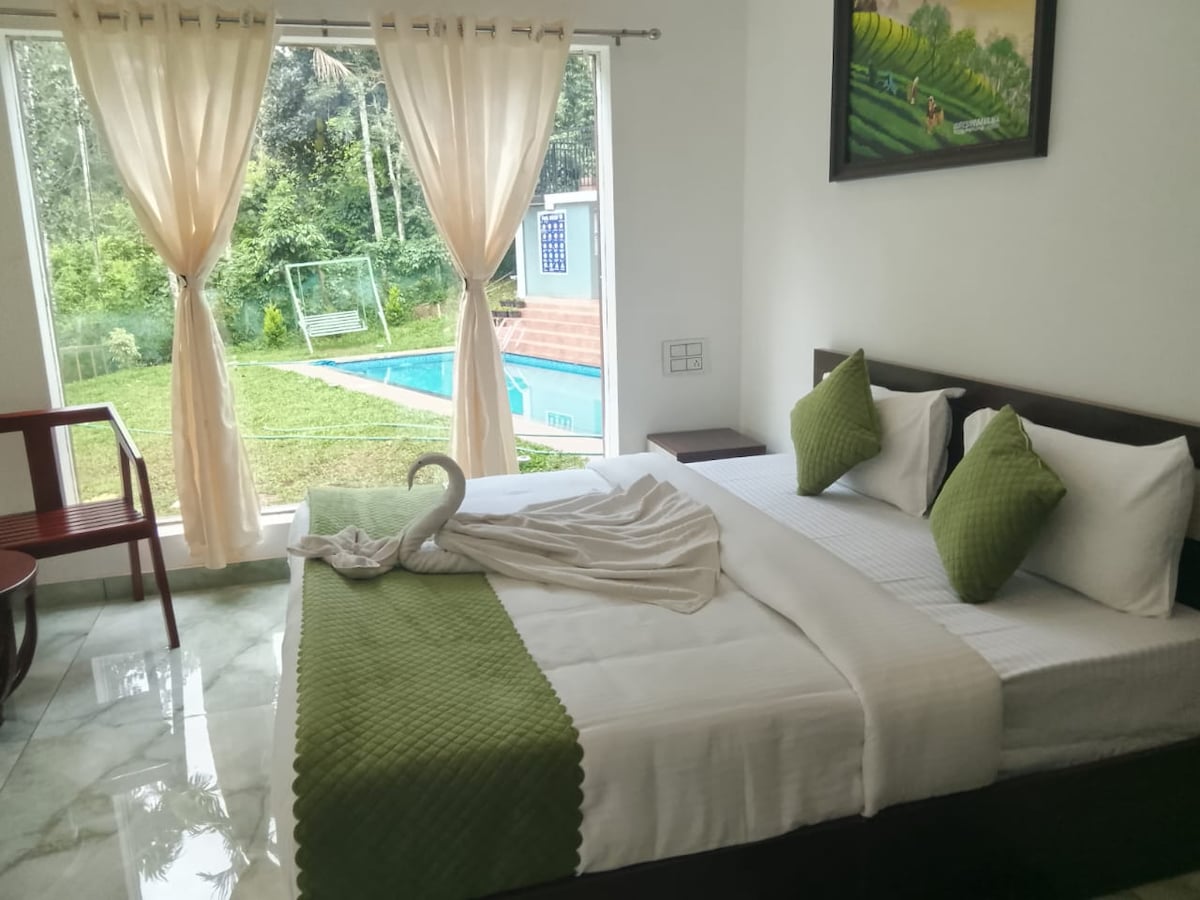 A bright bedroom is furnished with a spacious bed adorned with white and green accent pillows. Natural light enters through large windows, revealing a view of the outdoors and a turquoise pool. A wooden chair complements the serene setting.