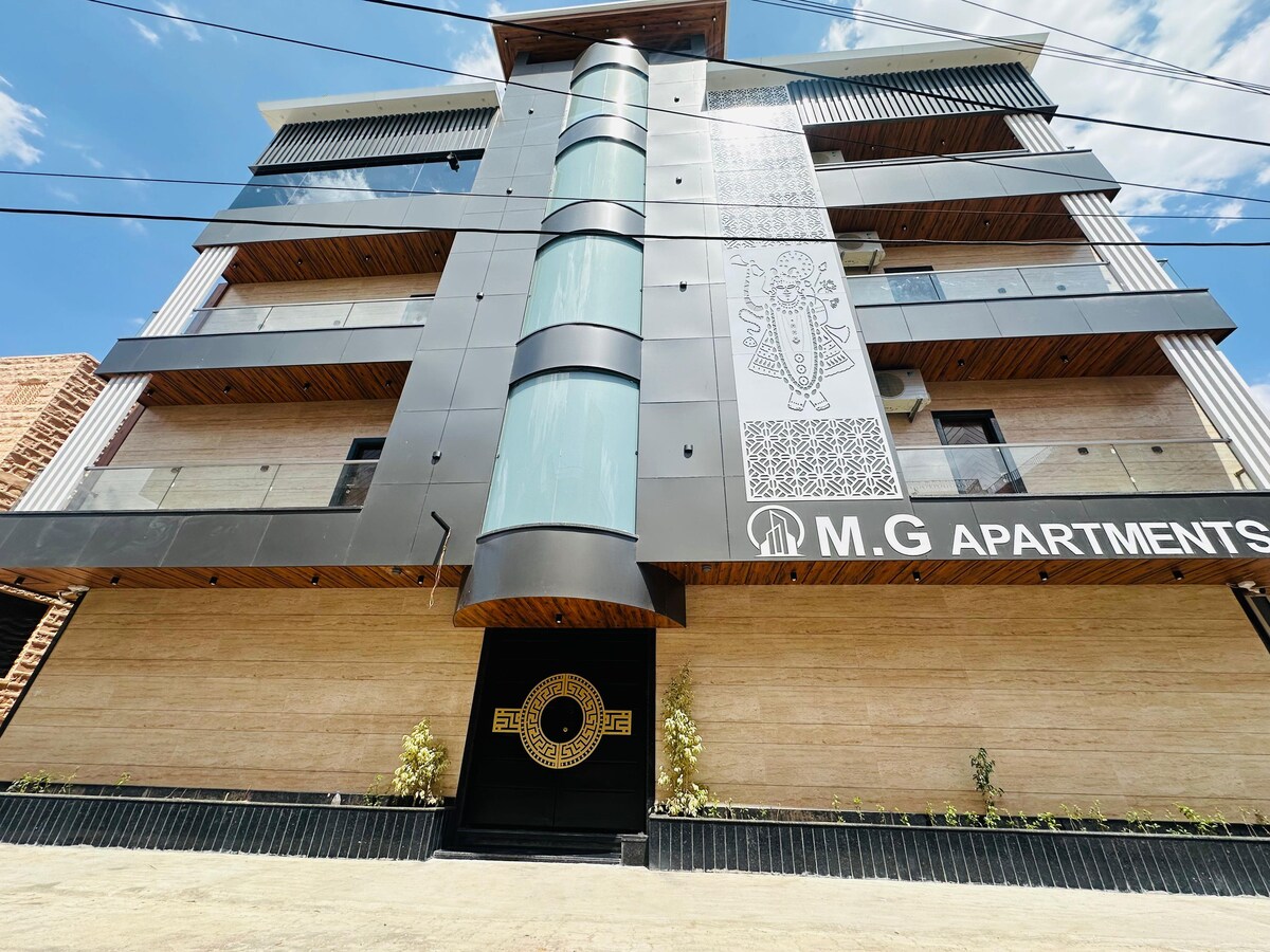 The exterior of a contemporary apartment building is presented, featuring a combination of sleek black and natural stone finishes. A large central column enhances the design, while balconies are visible on the upper floors. The entrance is marked by a distinctive logo and neatly arranged greenery.