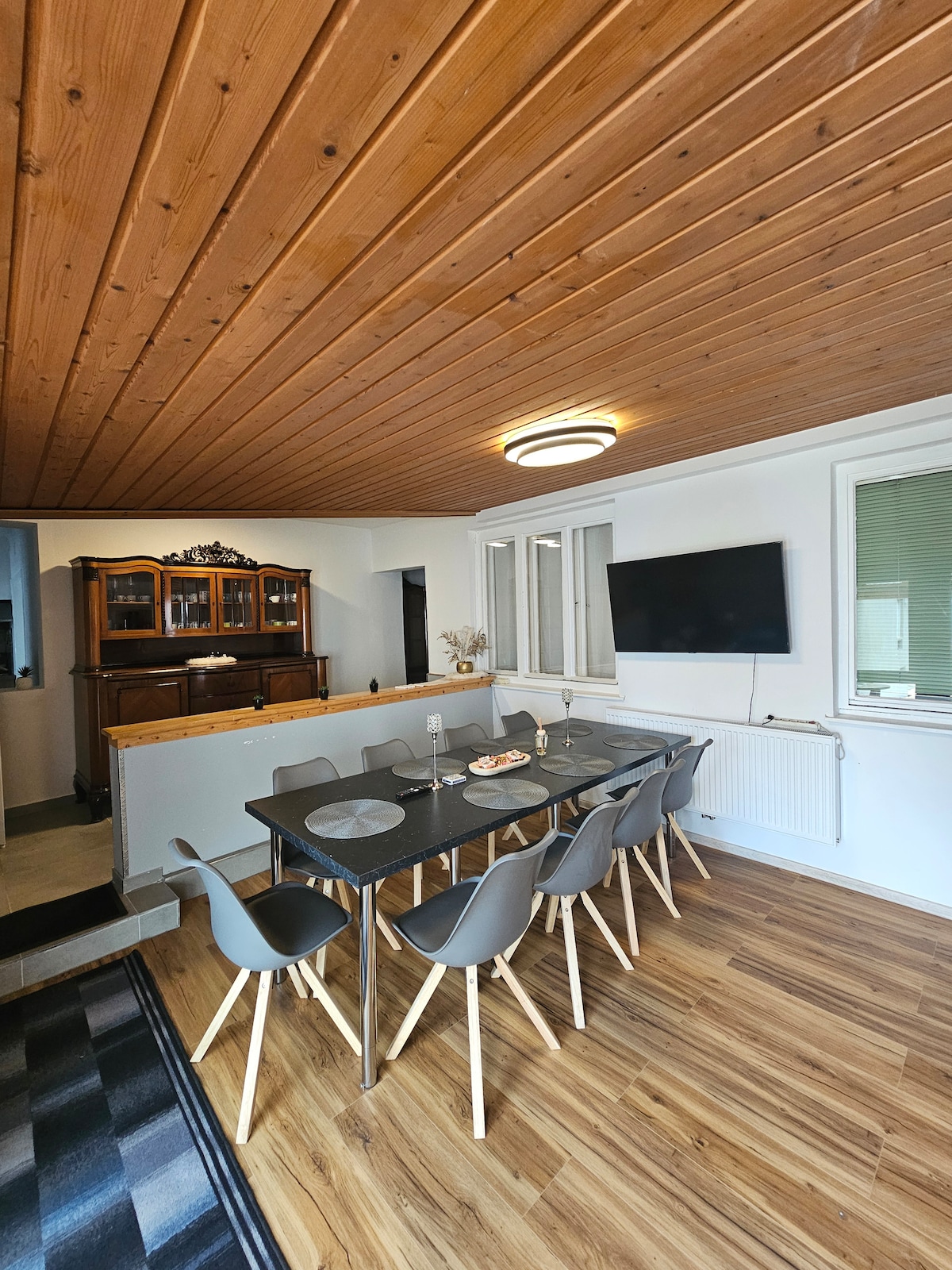 A dining area is presented with a large rectangular table surrounded by six chairs. Natural wood paneling covers the ceiling, while a wall-mounted television is visible nearby. A sideboard adds storage and display space, contributing to the room's functional layout.