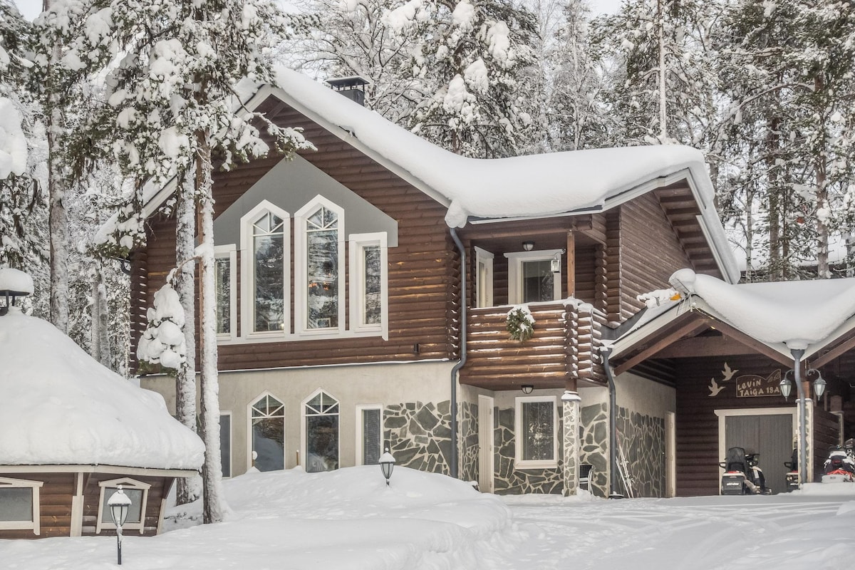 A two-story wooden cottage is surrounded by a snow-covered landscape, featuring large windows with geometric designs. The front entrance is accentuated by a stone façade, while a covered outdoor area is visible nearby. Snow-laden trees frame the peaceful setting, creating a serene winter atmosphere.
