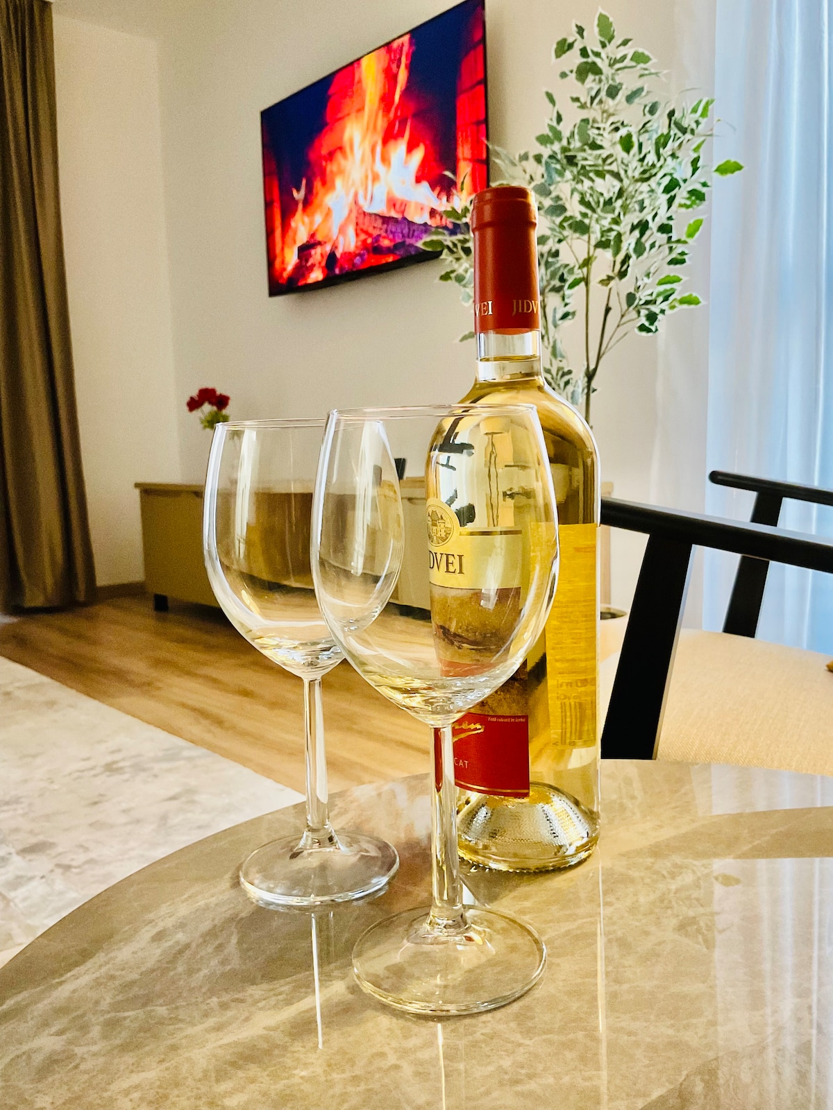 A dining table is set with two clear wine glasses and a bottle of white wine. In the background, a modern television displays a cozy fire, contributing to a warm ambiance. A decorative plant is positioned nearby, adding a touch of greenery.