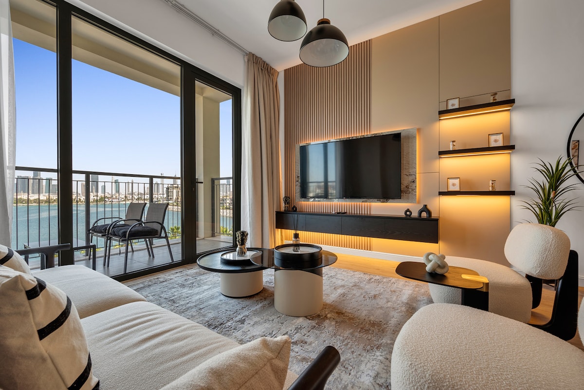 A modern living area features a large television mounted on a stylish wall unit, surrounded by shelf lighting. A comfortable seating arrangement includes a plush sofa and minimalist chairs. A balcony with glass doors provides a vibrant view of the sea and city skyline.