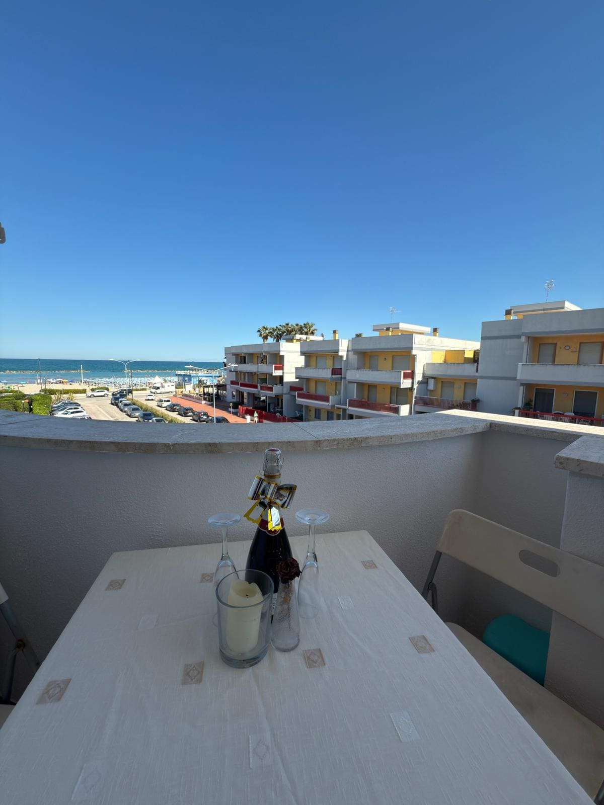 A balcony area features a small table set with glasses and a candle, overlooking a scenic view of the sea and nearby buildings. The clear blue sky enhances the inviting atmosphere of the outdoor space.