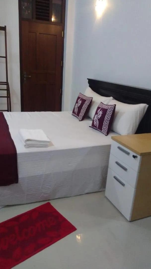 A comfortable room features a spacious bed with decorative pillows. A bedside table and a small storage unit are positioned nearby. The room's neutral colors are complemented by a welcome mat at the entrance. Soft lighting enhances the room's relaxing ambience.