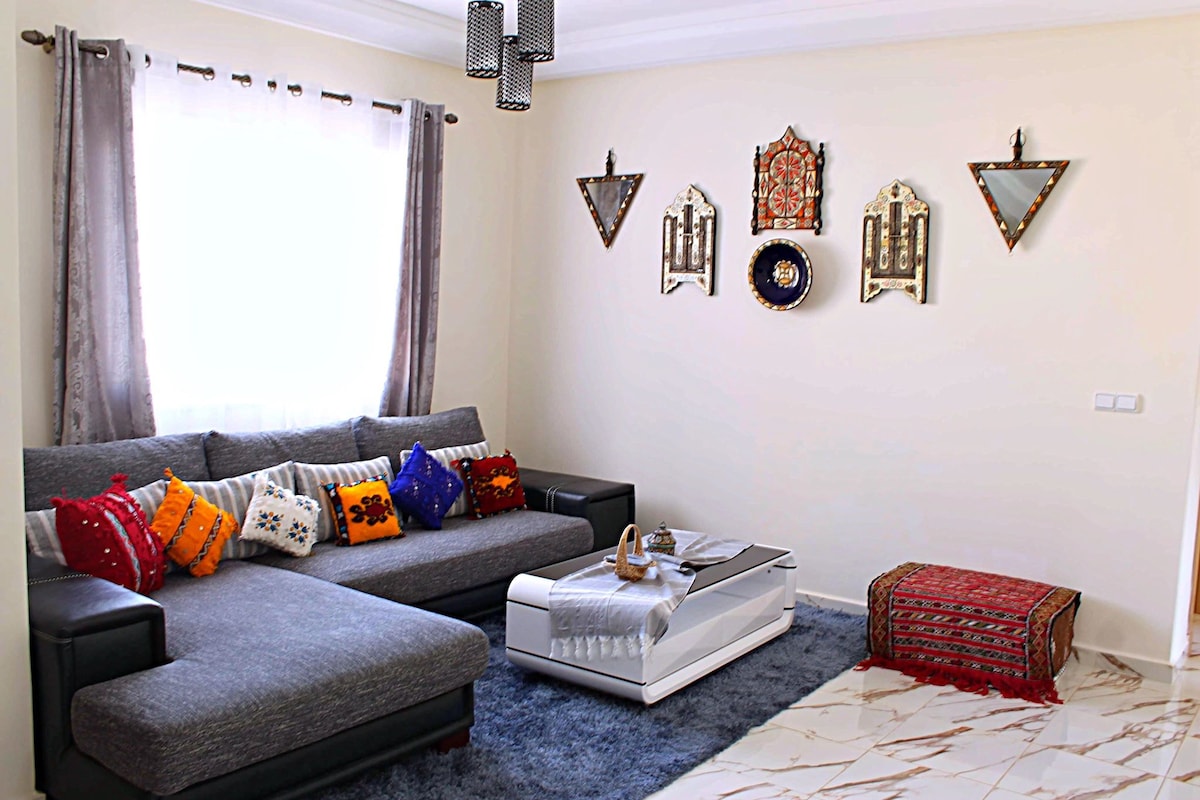 A spacious living area features a large sectional sofa adorned with colorful decorative cushions. A sleek coffee table sits in front, complemented by a textured area rug. The walls showcase an assortment of decorative art pieces, enhancing the room's contemporary character.