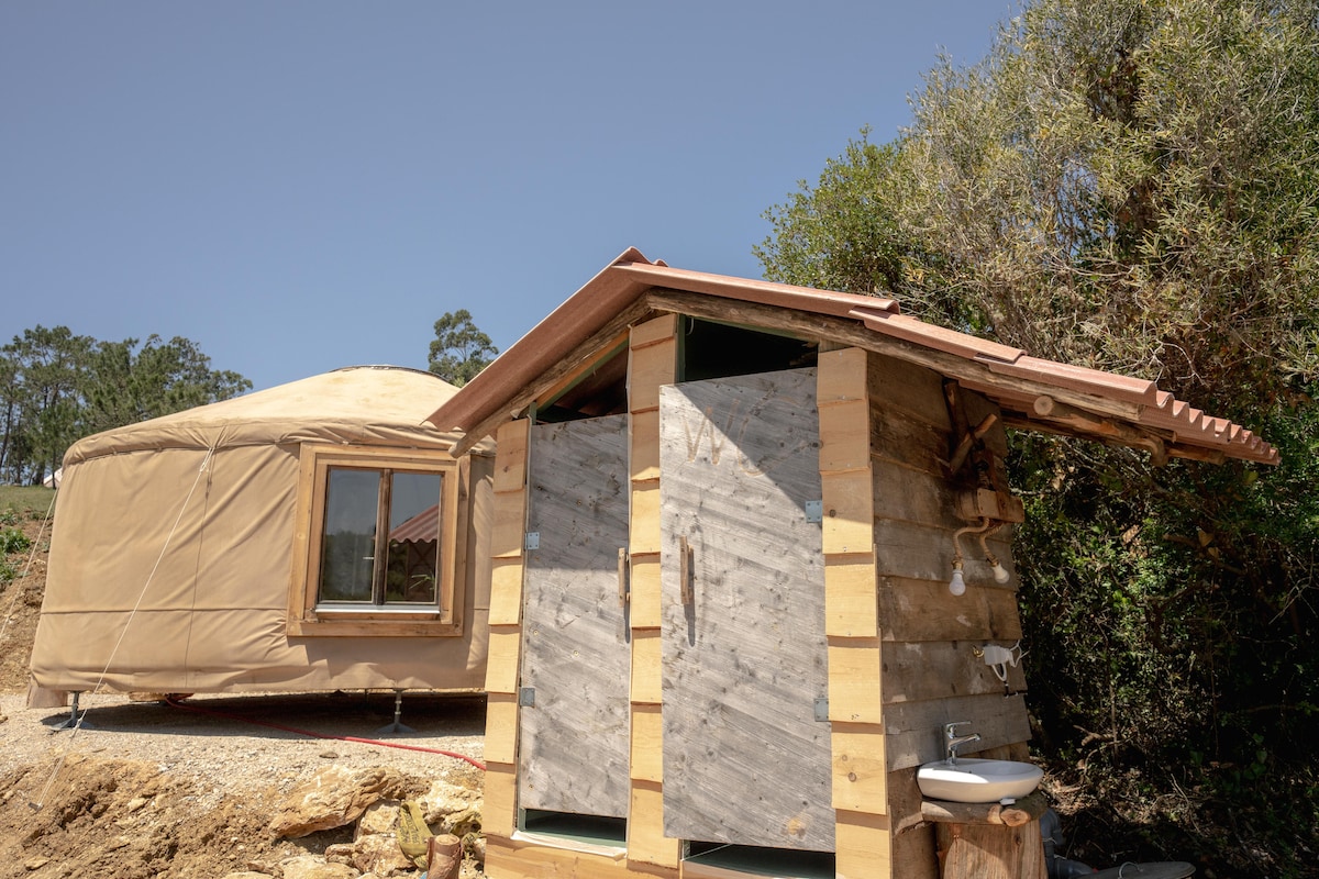 An outdoor view of a yurt beside a rustic wooden structure offering shared bathroom facilities. The yurt features a large window covered with a light fabric, allowing natural light to enter. The bathroom structure displays a simple sink and wooden doors, surrounded by greenery.