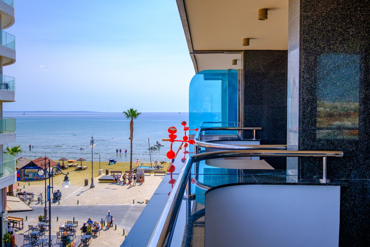 A balcony overlooks a sandy beach, with gentle waves lapping at the shore. Structures and palm trees line the beach, while people are seen enjoying the sun and water activities. The view includes clear skies and a distant horizon over the sea.