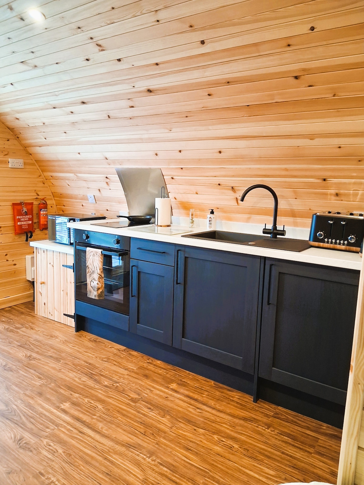 A modern kitchen area is presented, featuring wooden paneling and sleek cabinetry. A black faucet is positioned above a white countertop, which includes an oven and a compact coffee maker. Essential items like a kettle and utensils are arranged neatly, completing the functional space.